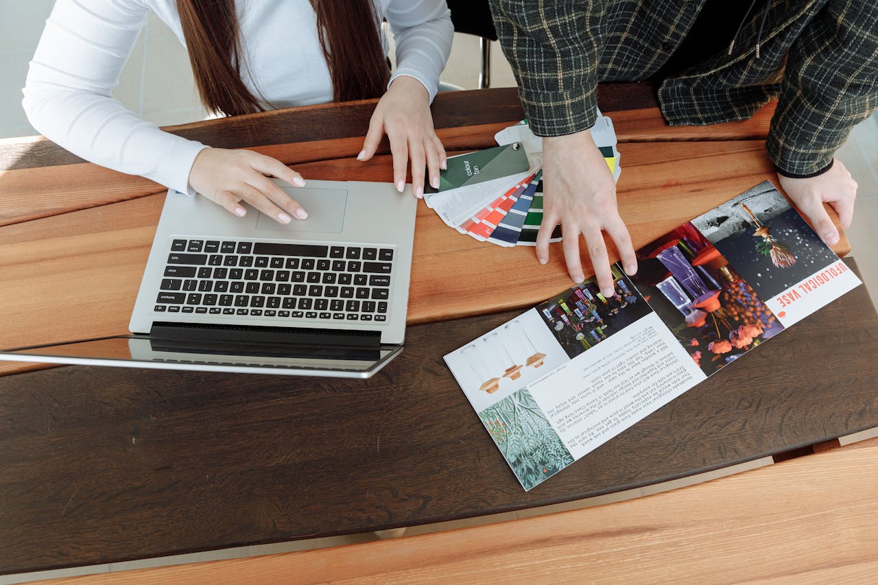 Two colleagues working on a design project using a laptop and color guides at a wooden table.